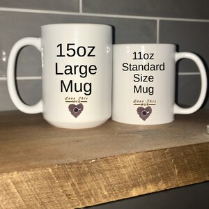 May include: Two white ceramic mugs with black text and a coffee bean heart design. The larger mug is labeled "15oz Large Mug" and the smaller mug is labeled "11oz Standard Size Mug".