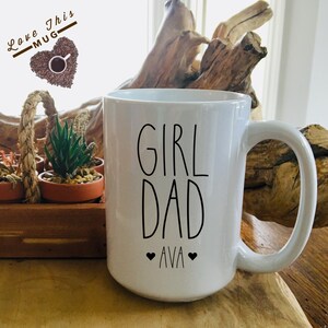 May include: White ceramic coffee mug with black text that reads "Girl Dad" and "Ava" with two small hearts on either side of the name.