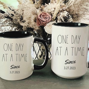 May include: Two white ceramic mugs with black rims and handles. Both mugs have the text "One Day At A Time" and the word "Since" followed by a date. The mug on the left has the date "3.27.2023". The mug on the right has the date "3.27.2023".