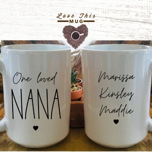 May include: Two white ceramic mugs with black text. One mug says "One loved NANA" with a small black heart. The other mug says "Marissa Kinsley Maddie" with a small black heart.
