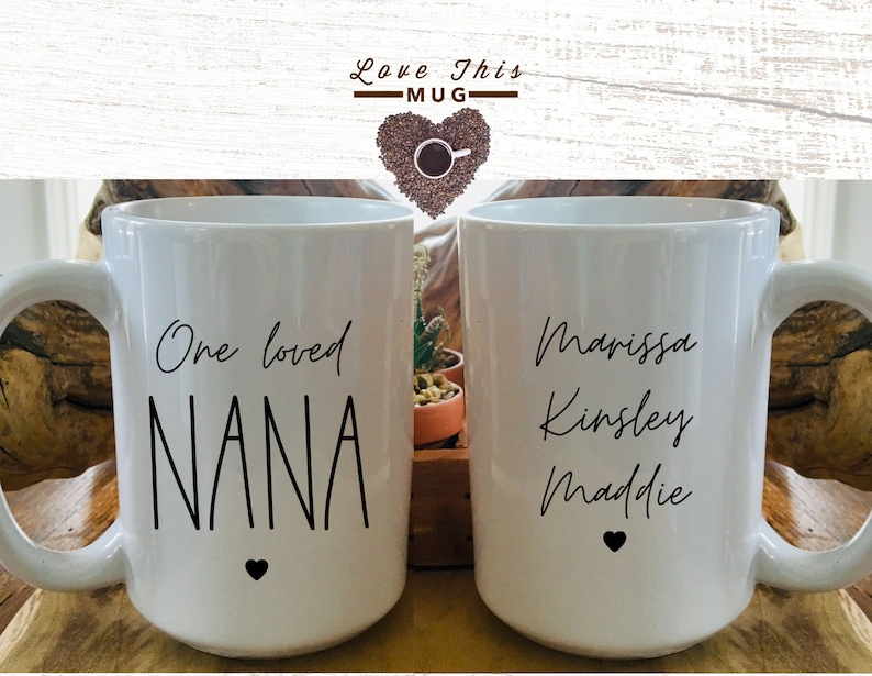 May include: Two white ceramic mugs with black text. One mug says "One loved NANA" with a small black heart. The other mug says "Marissa Kinsley Maddie" with a small black heart.