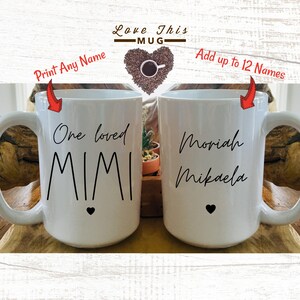 May include: Two white ceramic mugs with black handles. The mug on the left has the text "One loved Mimi" with a small black heart at the bottom. The mug on the right has the text "Moriah Mikaela".