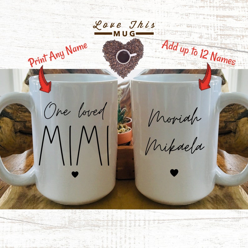 May include: Two white ceramic mugs with black handles. The mug on the left has the text "One loved Mimi" with a small black heart at the bottom. The mug on the right has the text "Moriah Mikaela".
