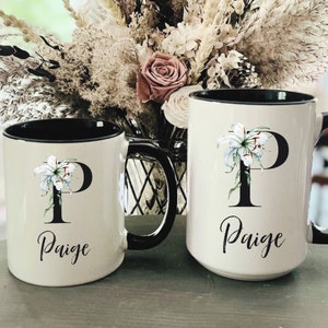 May include: Two white ceramic mugs with black rims and handles. Each mug features a large black letter "P" with a white lily and the name "Paige" in script. The mugs are different sizes.
