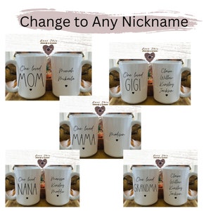 May include: Six white ceramic mugs with black text. The mugs are arranged in two rows of three. Each mug has a different nickname on it, such as "Mom", "Gigi", "Grandma", "Mama", "Nana", and a personalized name. The mugs are all decorated with a small black heart.