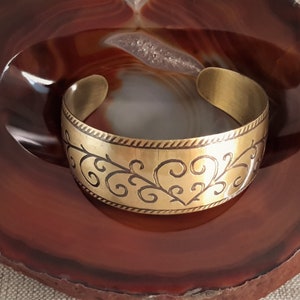 Könnte beinhalten: Ein goldfarbenes Metallarmband mit einem komplizierten floralen Design. Das Armband ist breit und hat eine abgerundete Form.