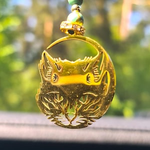 May include: A gold-colored cat pendant necklace with a detailed cat face design. The pendant hangs from a strand of green and gold beads. The necklace is displayed against a blurred green background.
