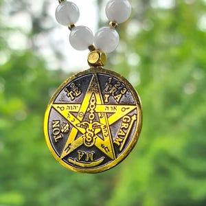 Puede incluir: Un collar con colgante que presenta un dije dorado en forma de estrella con símbolos y texto intrincados. El collar tiene una hebra de cuentas rojas y blancas. El fondo es verde borroso.