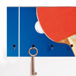 Peut inclure: Un porte-clés bleu sur le thème du ping-pong. Il présente une raquette rouge, une balle blanche et un fond bleu avec des lignes blanches. Une clé argentée est suspendue au porte-clés, et il y a trois crochets noirs.