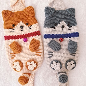 May include: Two crocheted cat-shaped bags with beige straps. One cat is brown and white with a red collar and the other is gray and white with a blue collar.