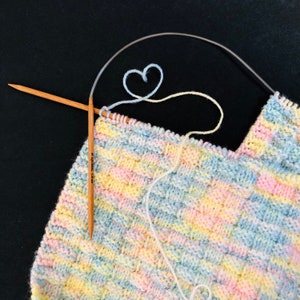 Puede incluir: Primer plano de una manta de bebé de punto de colores con una aguja de tejer de madera y un lazo de hilo blanco en forma de corazón.