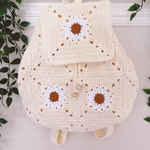 May include: A crocheted backpack with a white daisy pattern. The backpack has a drawstring closure and adjustable straps.