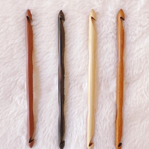 May include: Four wooden crochet hooks in varying shades of brown and tan. The hooks are arranged vertically against a white, textured background. Each hook has a curved end for catching yarn.