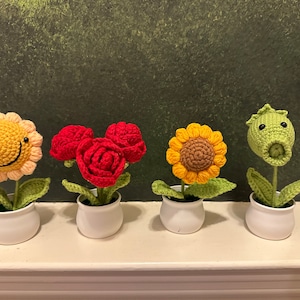 Puede incluir: Cuatro macetas de crochet con diferentes diseños de flores. Las macetas son blancas y las flores son amarillas, rojas y verdes.