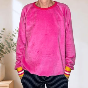 Könnte beinhalten: Ein langärmeliges Velours-Sweatshirt in leuchtendem Pink mit Rundhalsausschnitt und Raglanärmeln. Die Ärmelbündchen sind senfgelb und mit zwei horizontalen weißen Streifen versehen. Dazu wird eine dunkelblaue Hose mit feinen weißen Nadelstreifen getragen.