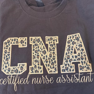 May include: A dark brown t-shirt with the embroidered text "CNA" in large, leopard print letters. Below, the words "certified nurse assistant" are embroidered in a cursive font. The shirt is made of a soft material.