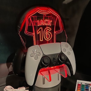 May include: A white and black PlayStation 5 controller is on a stand with a red light-up jersey with the name "JACK" and the number "16" on it.