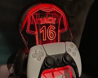 May include: A white and black PlayStation 5 controller is on a stand with a red light-up jersey with the name "JACK" and the number "16" on it.