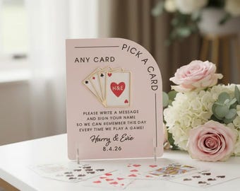 Puede incluir: Un letrero de tarjeta de boda rosa con el texto "Pick a Card" y "Any Card". El letrero presenta un diseño de naipe con las iniciales "H&E" dentro de un corazón. El letrero también incluye los nombres "Harry & Evie" y la fecha "8.4.26".