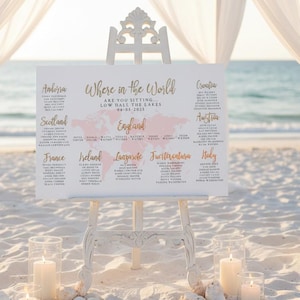 May include: A wedding seating plan on a white easel, with the words "Where in the World" and a world map. The sign is on a sandy beach, with candles and seashells in front. Sheer white fabric drapes in the background, with the sea visible.