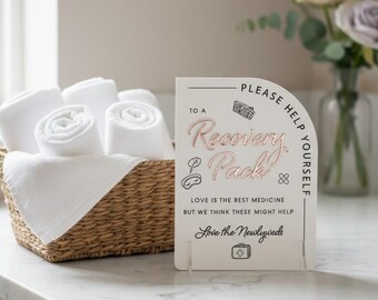 May include: A woven basket filled with rolled white towels sits next to a sign that reads "Recovery Pack". The sign has text that says "Love is the best medicine but we think these might help". A vase of flowers is in the background.