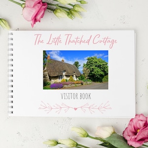 May include: A white visitor book with a pink title that reads "The Little Thatched Cottage" and a photo of a thatched cottage. The book has a spiral binding and a pink floral design at the bottom.