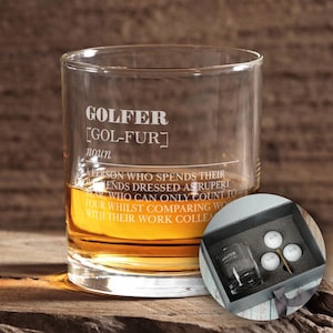 May include: A clear glass whisky tumbler with the word "GOLFER" etched on the side, along with the definition "[GOL-FUR] noun A PERSON WHO SPENDS THEIR WEEKENDS DRESSED AS RUPERT BEAR WHO CAN ONLY COUNT TO FOUR WHILST COMPARING WORK WITH THEIR WORK COLLEAGUES". The tumbler is filled with amber liquid and is sitting on a wooden surface. A black box with a white golf ball and a gold golf tee is in the background.