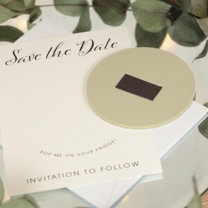 May include: A white card with the text "Save the Date" in black cursive writing. Below the text is the phrase "Pop me on your fridge!" and "Invitation to follow". A beige round magnet with a brown rectangular magnet on top is on the card.