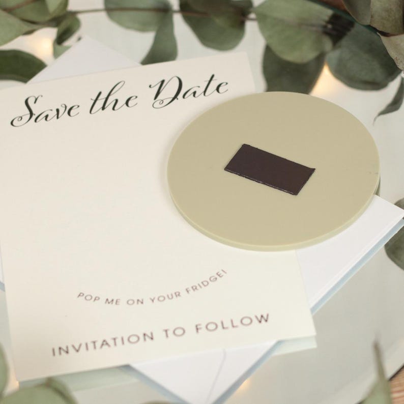 May include: A white card with the text "Save the Date" in black cursive writing. Below the text is the phrase "Pop me on your fridge!" and "Invitation to follow". A beige round magnet with a brown rectangular magnet on top is on the card.