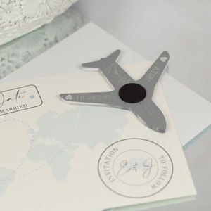 May include: Silver airplane shaped wedding favor with a magnet on the back. The airplane has the text "IBIZA" and "10.02.2023" engraved on it. The favor is on a white card with a blue world map and the text "Date Married" and "Invitation to follow".