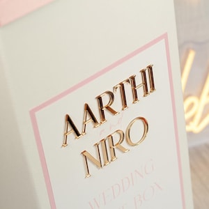 May include: A white rectangular box featuring a light pink border and gold-colored raised lettering that reads AARTHI and NIRO with the word and in script. Below the names, the text WEDDING GUEST BOX is printed in a light pink sans-serif font.