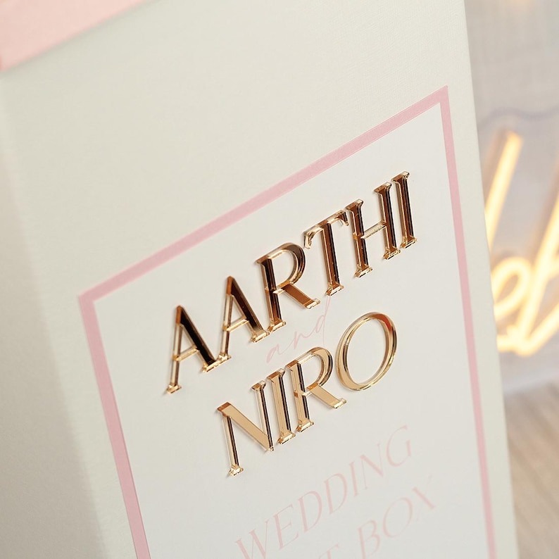 May include: A white rectangular box featuring a light pink border and gold-colored raised lettering that reads AARTHI and NIRO with the word and in script. Below the names, the text WEDDING GUEST BOX is printed in a light pink sans-serif font.