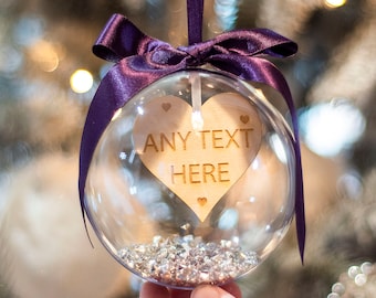 May include: A clear spherical Christmas ornament containing small silver crystals and a wooden heart cutout. The heart is engraved with the text ANY TEXT HERE and three small hearts. A dark purple satin ribbon is tied at the top of the ornament.