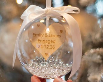 May include: A clear spherical ornament filled with small clear crystals. Inside hangs a wooden heart engraved with the text Emily & James Engaged 24.12.26. A white satin ribbon is tied in a bow at the top of the ornament for hanging purposes.