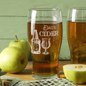 May include: Two clear glass mugs filled with amber colored cider. The mug on the left has the text "Dad's Cider" etched on the side, along with a bottle and a glass of cider.
