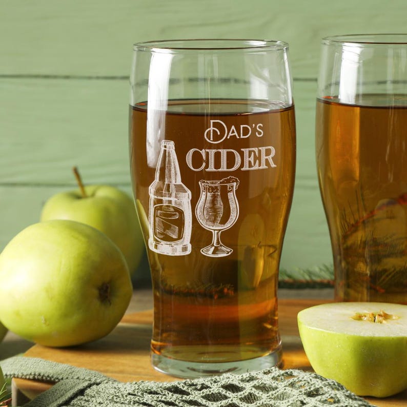 May include: Two clear glass mugs filled with amber colored cider. The mug on the left has the text "Dad's Cider" etched on the side, along with a bottle and a glass of cider.