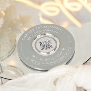 May include: A circular, grey disc with the inscription "CAPTURE THE MEMORIES MR & MRS JOHNSON 23.07.2026" and "SCAN THE QR CODE TO UPLOAD YOUR SNAPS FROM THE DAY". A QR code is in the centre. The disc is on a reflective surface.