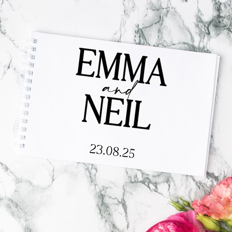 May include: A white spiral-bound notebook with black text that reads EMMA and NEIL followed by the date 23.08.25. The cover features a clean, minimalist design with bold serif lettering for the names and a script font for the word and.