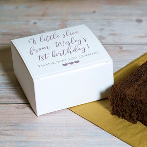 May include: A white box with a purple inscription that reads "A little slice from Wesley's 1st birthday! Thank you for coming" with three purple hearts. The box is sitting on a wooden surface next to a brown chocolate cake.