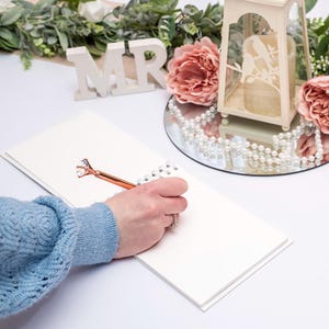May include: A white spiral-bound guest book with a rose gold pen featuring a large clear diamond-shaped topper. The pen is positioned on the blank page of the notebook. Nearby are white wooden letters spelling MR, pink flowers, and a lantern.