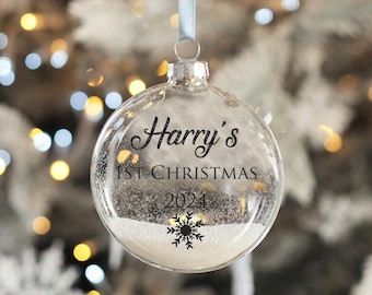 以下が含まれることがあります： 雪の結晶のデザインと「Harry's 1st Christmas 2024」の文字が書かれた透明なガラス製のクリスマスオーナメント