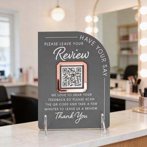 May include: A grey sign with a rose gold frame around a QR code, encouraging customers to leave a review. The sign reads "PLEASE LEAVE YOUR Review" and "WE LOVE TO HEAR YOUR FEEDBACK". The sign is propped up on a white surface.