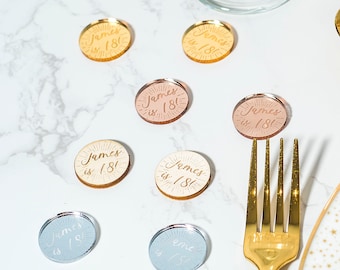 May include: Nine round, gold, silver, and pink acrylic place cards with the text "James is 18!" written in gold calligraphy. The place cards are scattered on a white marble surface with a gold fork in the foreground.