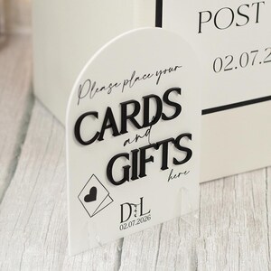 May include: An arched white acrylic sign featuring raised black lettering that reads "Please place your CARDS and GIFTS here". The sign includes a black envelope icon with a heart, the initials "DL", and the date "02.07.2026" printed in black.