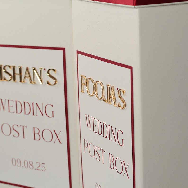 May include: Two off-white rectangular wedding post boxes featuring a deep red border. The boxes display raised gold lettering reading "ISHAN'S WEDDING POST BOX" and "POOJA'S WEDDING POST BOX" with the date "09.08.25" printed in red below the text.