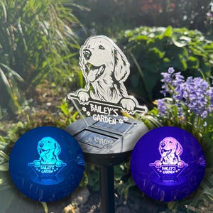 May include: A solar powered garden light with a clear acrylic panel featuring a silhouette of a golden retriever dog with the text "Bailey's Garden". The light is illuminated in blue and purple.