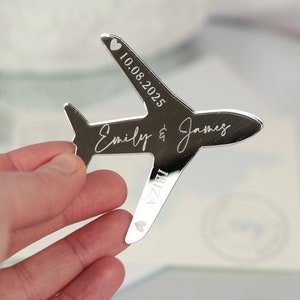 May include: Silver airplane shaped mirror with the text "Emily & James" and "IBIZA" engraved on it. The date "10.08.2025" is also engraved on the airplane.