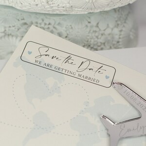 May include: A white card with a black rectangle that says "Save the Date" with a small blue heart on either side. The text below the rectangle says "We are getting married". The card has a light blue map of the world in the background.