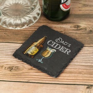 May include: A black slate coaster with the text "Dad's Cider" in white lettering. A drawing of a bottle of cider and a glass of cider are on the coaster.
