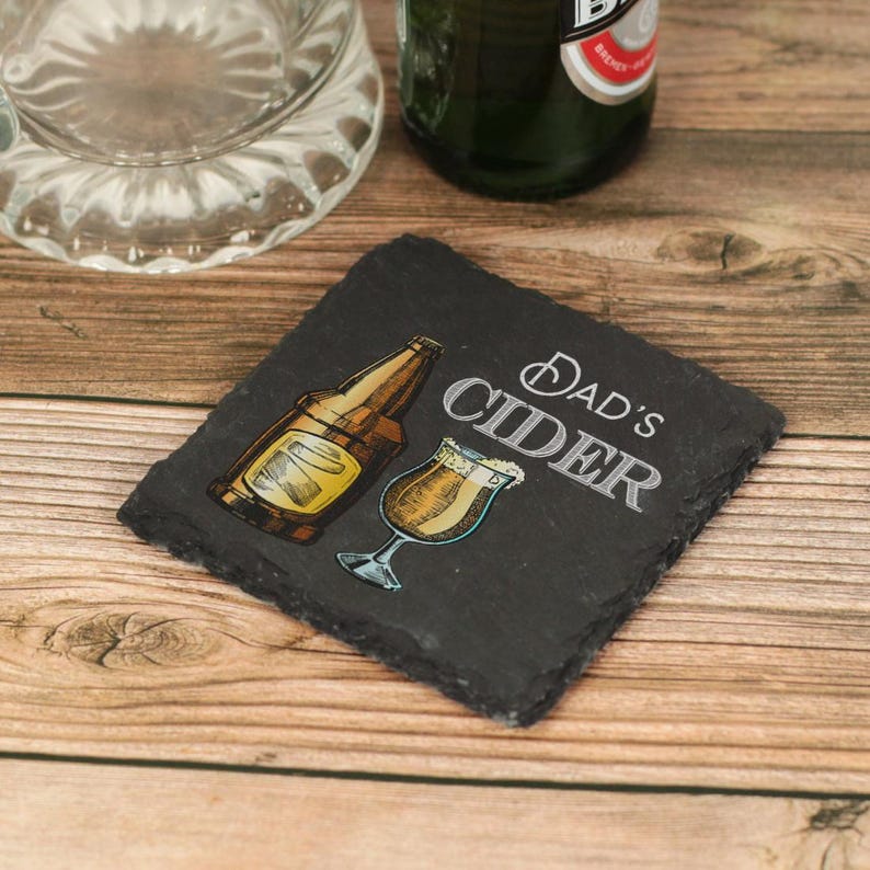 May include: A black slate coaster with the text "Dad's Cider" in white lettering. A drawing of a bottle of cider and a glass of cider are on the coaster.